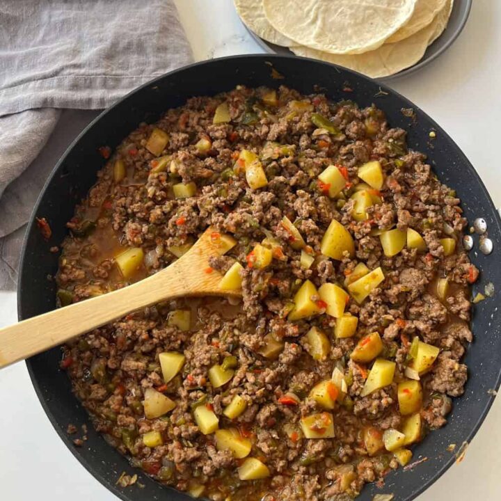 There is a large dark skillet with a ground beef and potato mixture in it. There is a wooden spoon resting in the skillet. In the background there is a gray plate with round corn tortillas on it and a gray cloth napkin. Items are on a white surface.