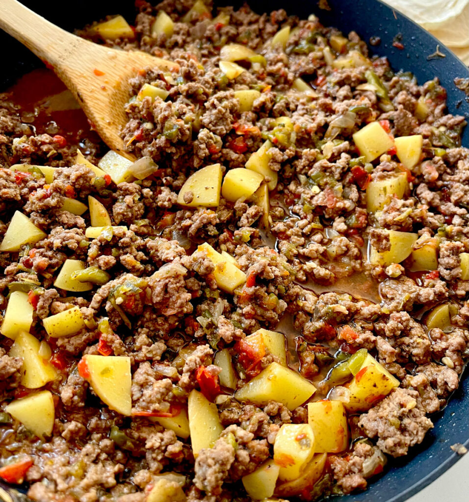 There is a large dark skillet with a ground beef and potato mixture in it. There is a wooden spoon resting in the skillet.