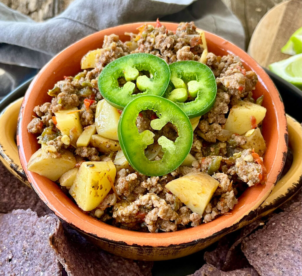 There is a orange bowl with a ground beer and potato mixture in it. There are sliced jalapeno peppers on top of the mixture. The bowl is on a large black shallow round dish. There are blue corn chips on the dish. There is a gray cloth napkin. Items are on a wood surface.