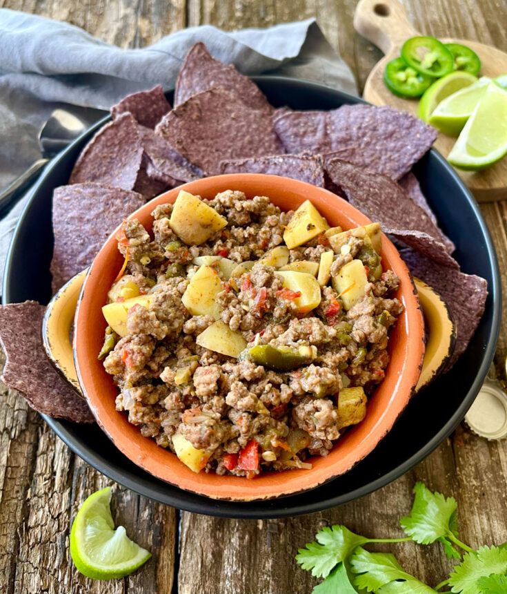 There is a orange bowl with a ground beer and potato mixture in it. The bowl is on a large black shallow round dish. There are blue corn chips on the dish. There are lime wedges and sliced green jalapeno peppers on a round wood plate and a gray cloth napkin. Items are on a wood surface.