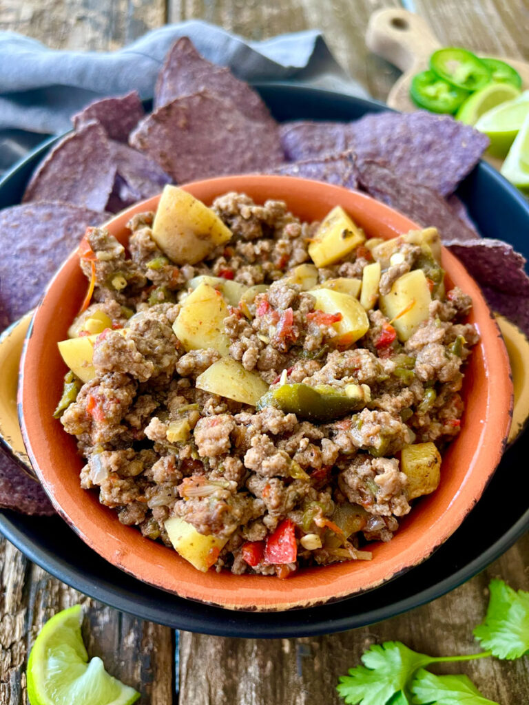 There is a orange bowl with a ground beer and potato mixture in it. The bowl is on a large black shallow round dish. There are blue corn chips on the dish. There are lime wedges and a gray cloth napkin. Items are on a wood surface.