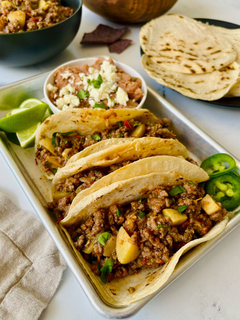 There is a metal tray with three round corn tortillas filled with a ground beef and potato mixture. There is a small white bowl on the tray with beans and cheese in it. There are lime wedges and sliced green jalapeno peppers on the tray. In the background there is a black plate with round corn tortillas on it. a black bowl with a ground beef and potato mixture in it. Item are on a white surface.