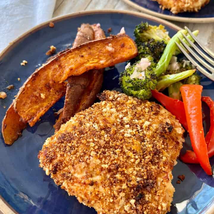 There is a blue plate with a piece of baked chicken breast with bread crumbs, slices of sweet potato, pieces of broccoli, and slices of red pepper. There is a fork resting on the plate. There is a white cloth napkin. Items are on a wood surface.