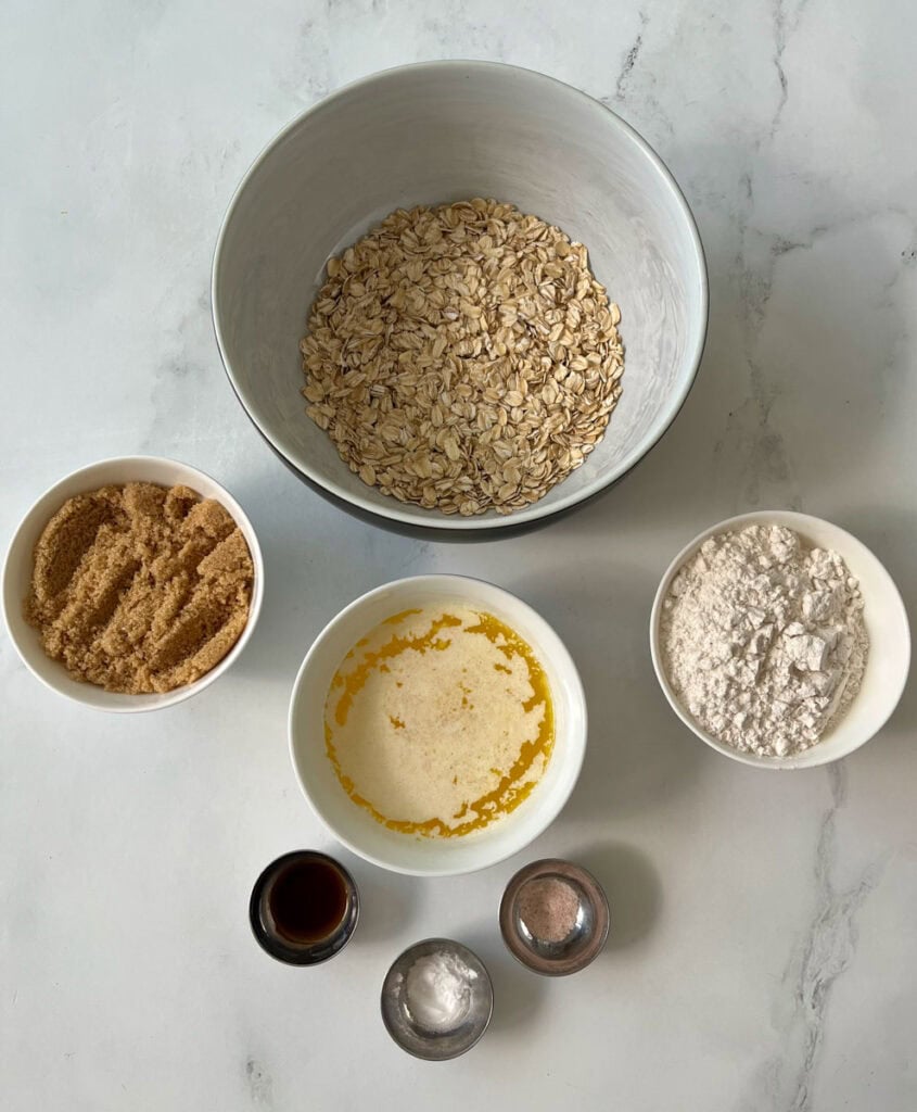 There is a large bowl with oats in it. There is a white bowl with brown sugar, a bowl with melted butter, and a bowl with flour in it. There are three very small bowls, one with vanilla extract, one with white baking soda, and one with salt. Items are on a white surface.