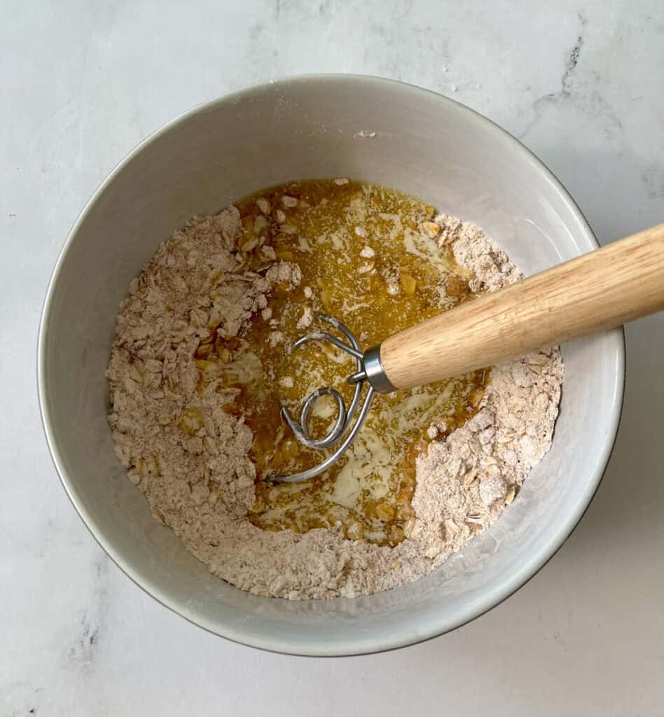 There is a large mixing bowl with flour, oats, brown sugar, and melted butter in it.There is a wood handle mixing tool resting in the bowl. Bowl is on a white surface.