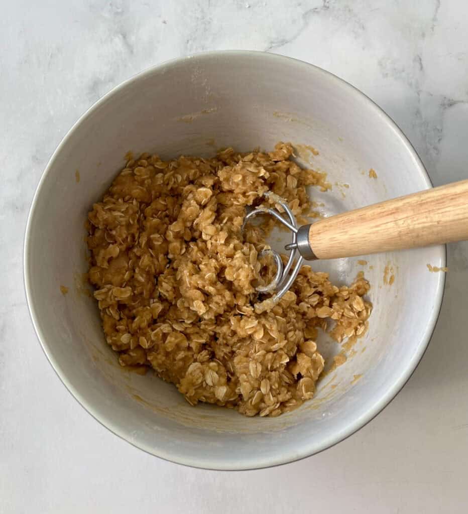 There is a large white bowl with a crumbly oatmeal mixture in it. There is a wood handle mixing tool resting in the bowl. Bowl is on a white surface.