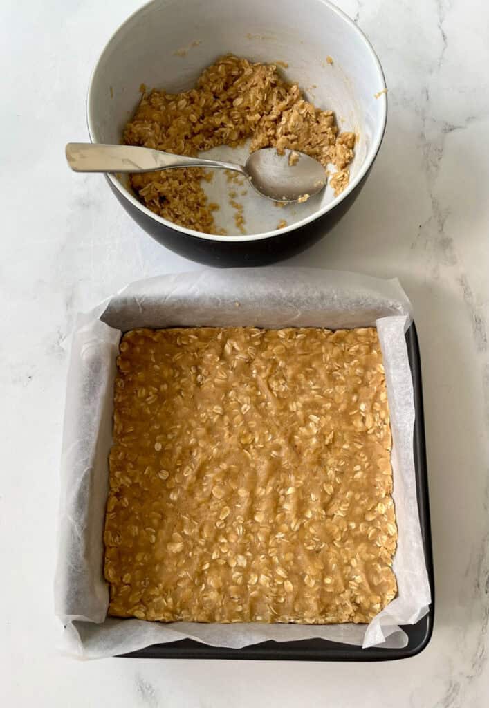 There is a square baking pan with a layer of an oatmeal mixture in it. There is a large bowl with a crumbly oatmeal mixture in it. There is a spoon resting in the bowl. Items are on a white surface.