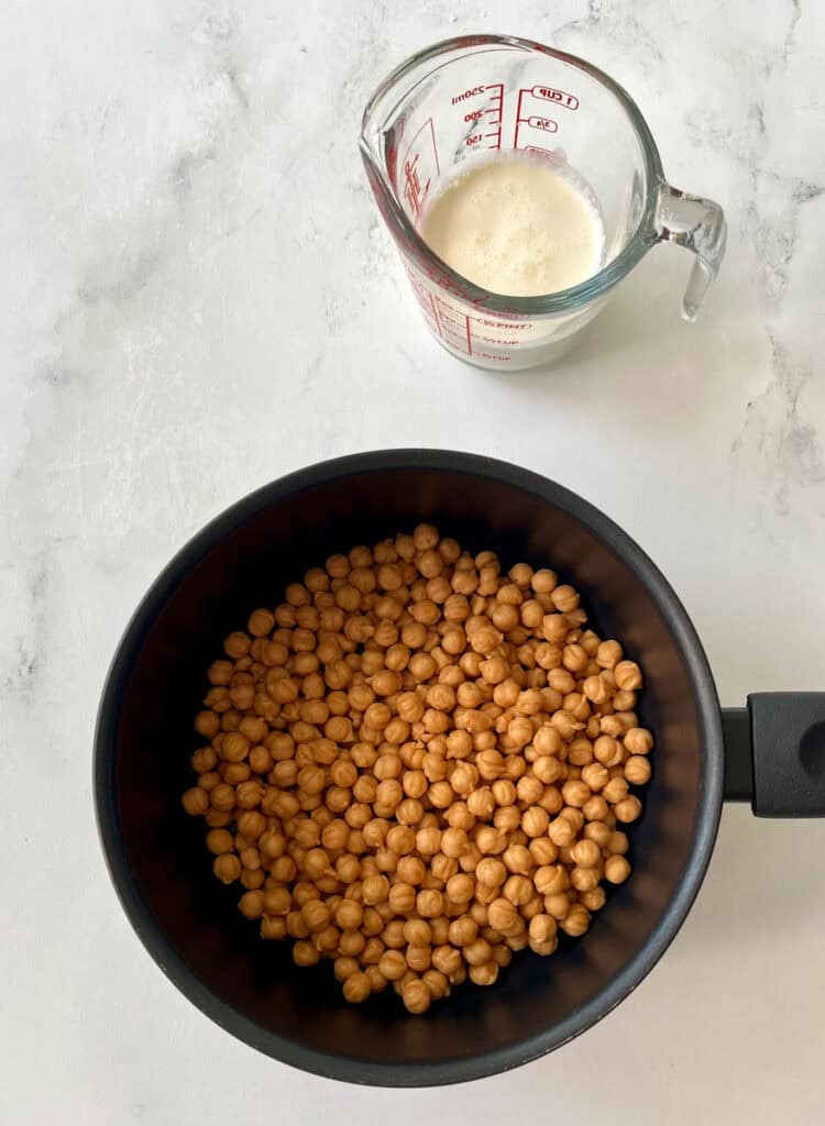 There is a dark saucepan with small round balls of caramel in it. There is a glass measuring cup with heavy cream in it. Items are on a white surface.