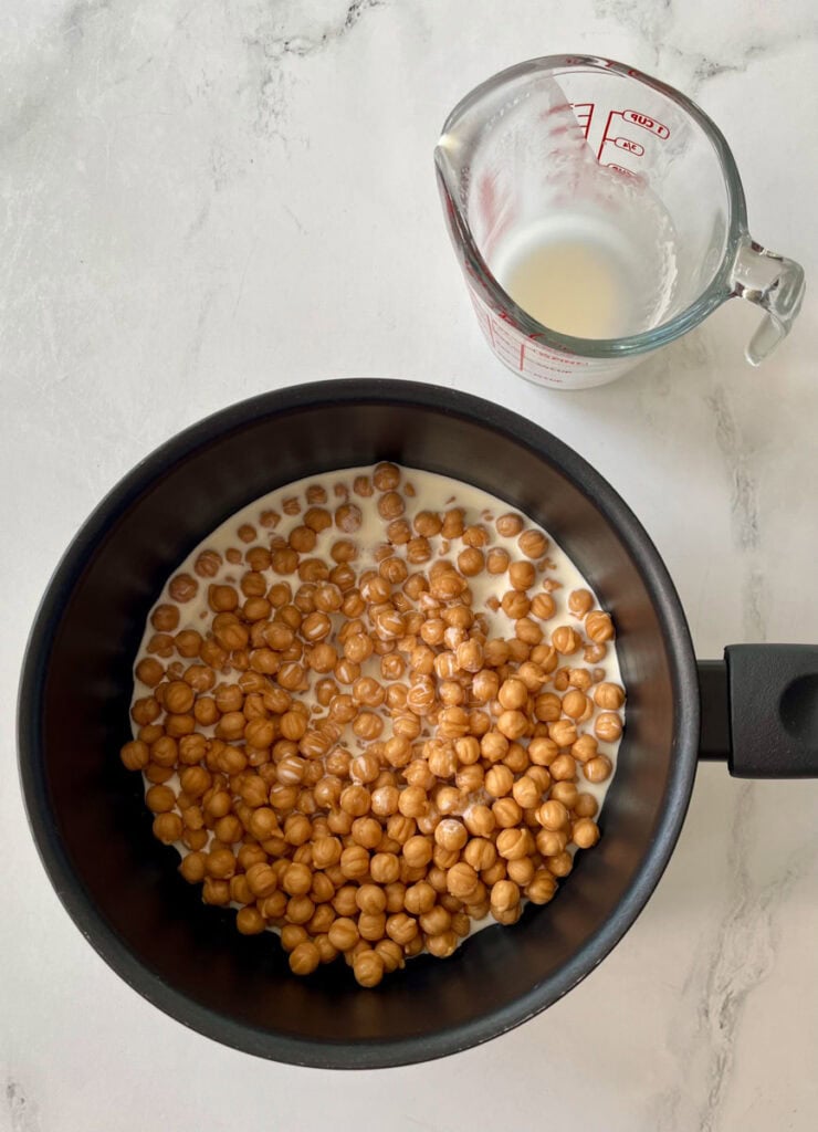 There is a dark saucepan with small round balls of caramel and heavy cream in it. There is an empty glass measuring cup. Items are on a white surface.