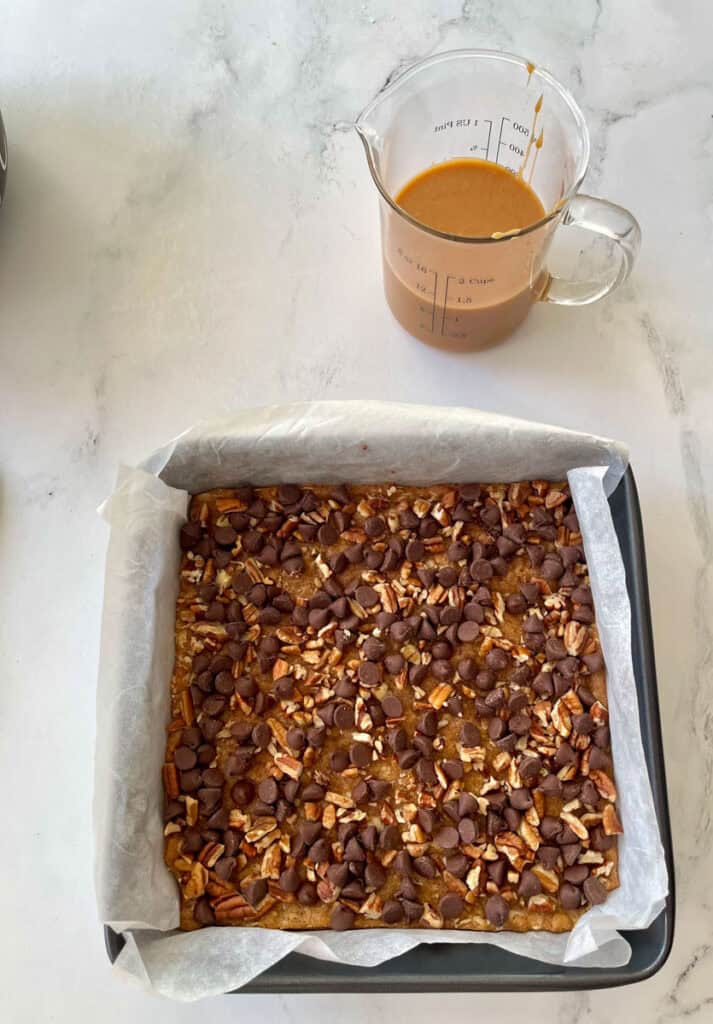 There is a square baking pan with a layer of an oatmeal mixture, a layer of chocolate chips and chopped nuts. There is a clear glass measuring cups with caramel sauce in it. Items are on a white surface.