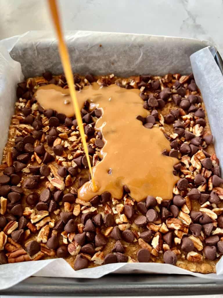 There is a square baking pan with a layer of an oatmeal mixture, a layer of chocolate chips and chopped nuts. There is caramel sauce being poured over the top of the layers.