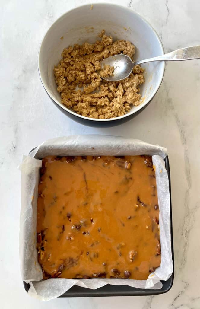 There is a square baking pan lined with white paper. There is an unbaked layered oatmeal mixture with caramel sauce poured over it. There is a large bowl with a crumbly oatmeal mixture in the background. There is a spoon resting in the bowl. Items are on a white surface.