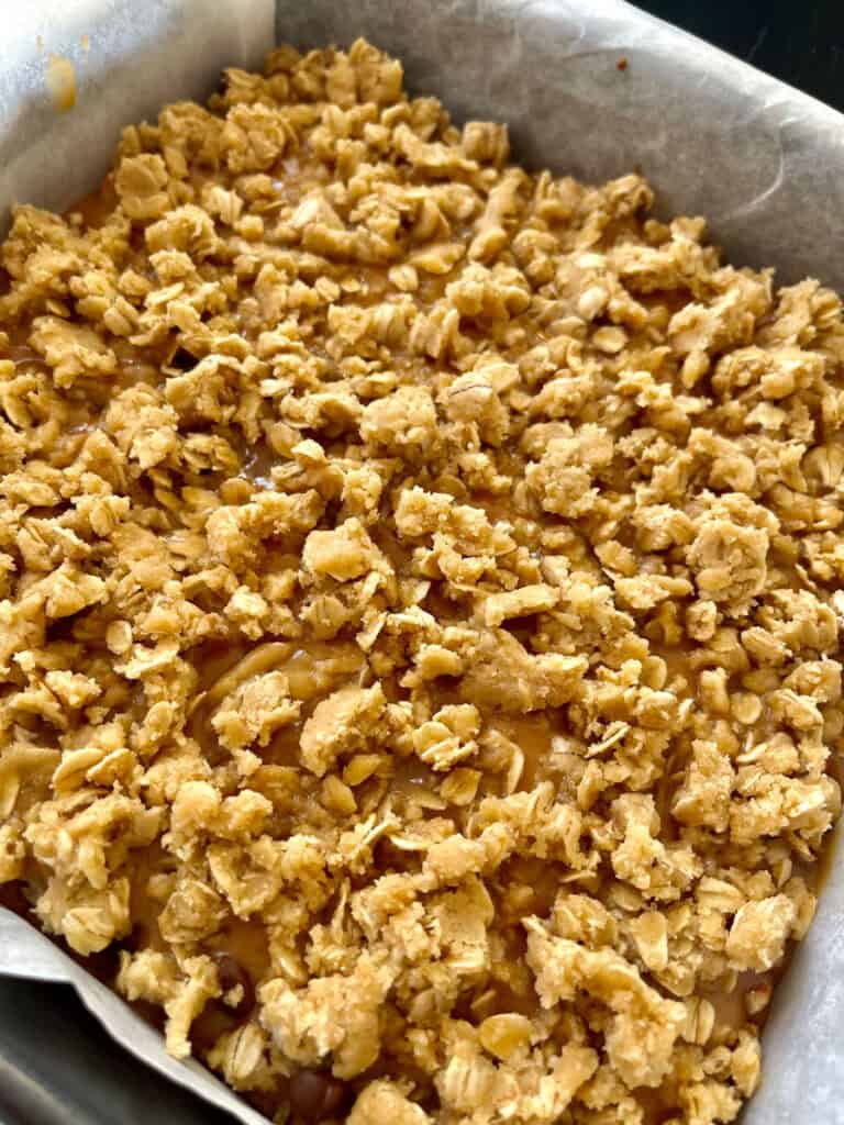 There is a square baking pan lined with white paper. There is an unbaked layered oatmeal mixture in it.