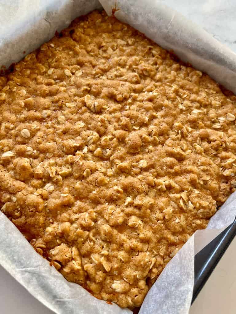 There is a square baking pan lined with white paper. There is a baked layered oatmeal mixture in it.