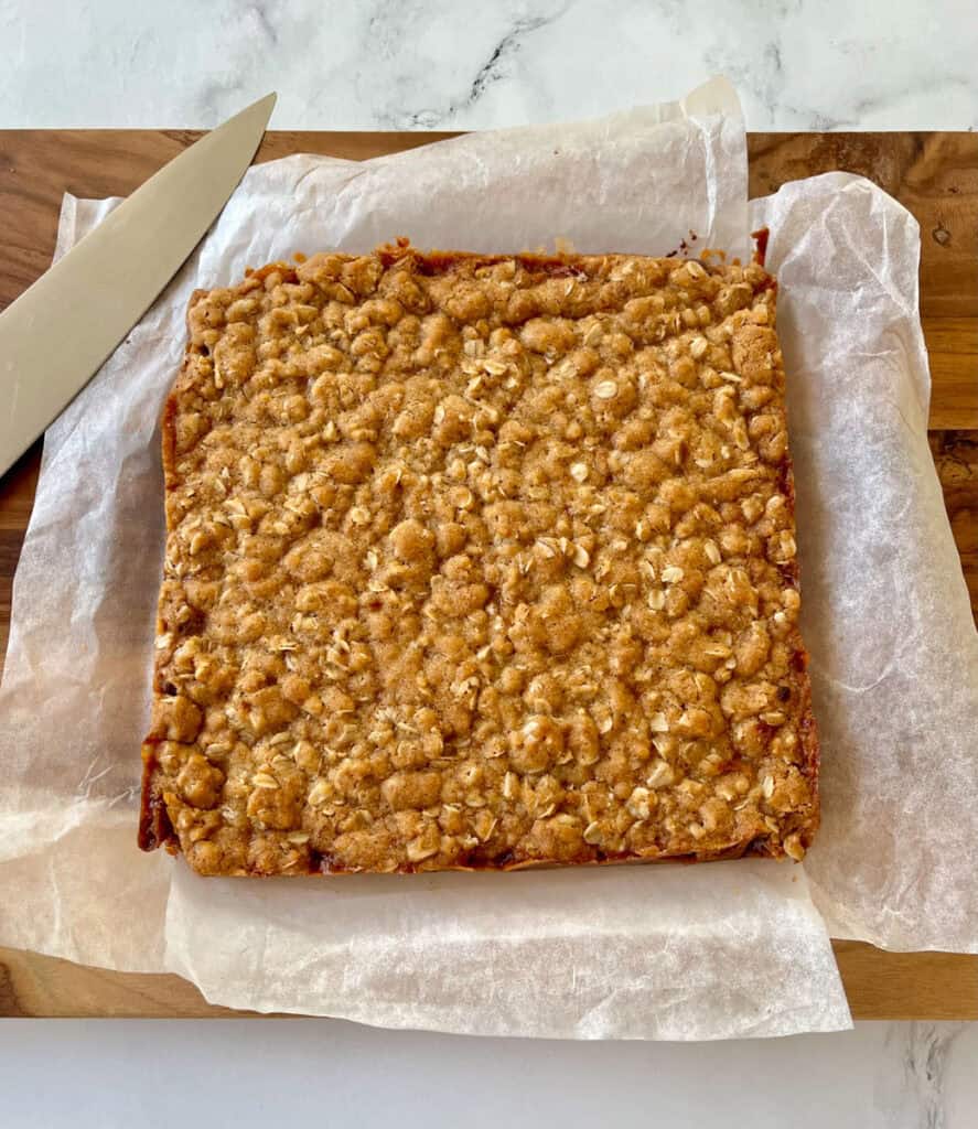 There is a large uncut square of baked oatmeal bar cookies on white paper on a wood cutting board. There is a large knife on the board. Wooden board is on a white surface.