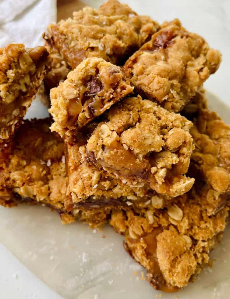 There is a pile of square layered bar cookies made with oatmeal, caramel, and chocolate chips. Bars are on a marble surface.