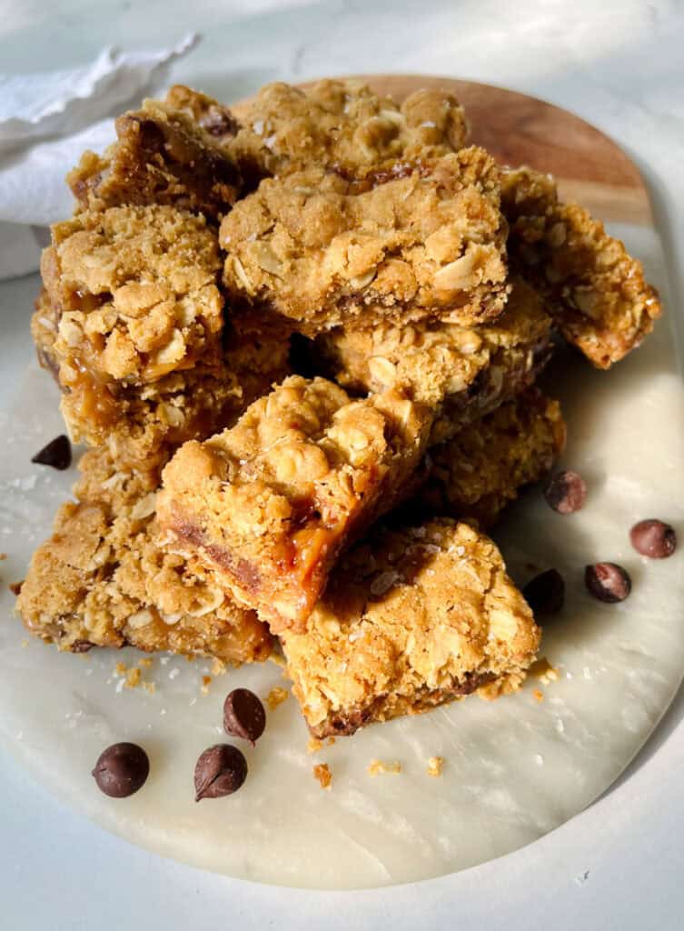 There is a pile of square layered bar cookies made with oatmeal, caramel, and chocolate chips. Bars are on a marble board. There are chocolate chips on the board. There is a white cloth napkin. Items are on a white surface.