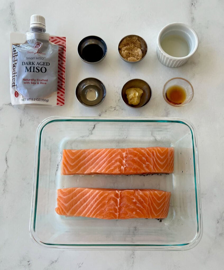 There is a rectangular clear glass dish with two salmon filets. There is a pouch container with Miso paste in it. There are multiple small dishes with ingredients in them: soy sauce, minced ginger, brown sugar, oil, and Asian cooking wine. Items are on a white surface.