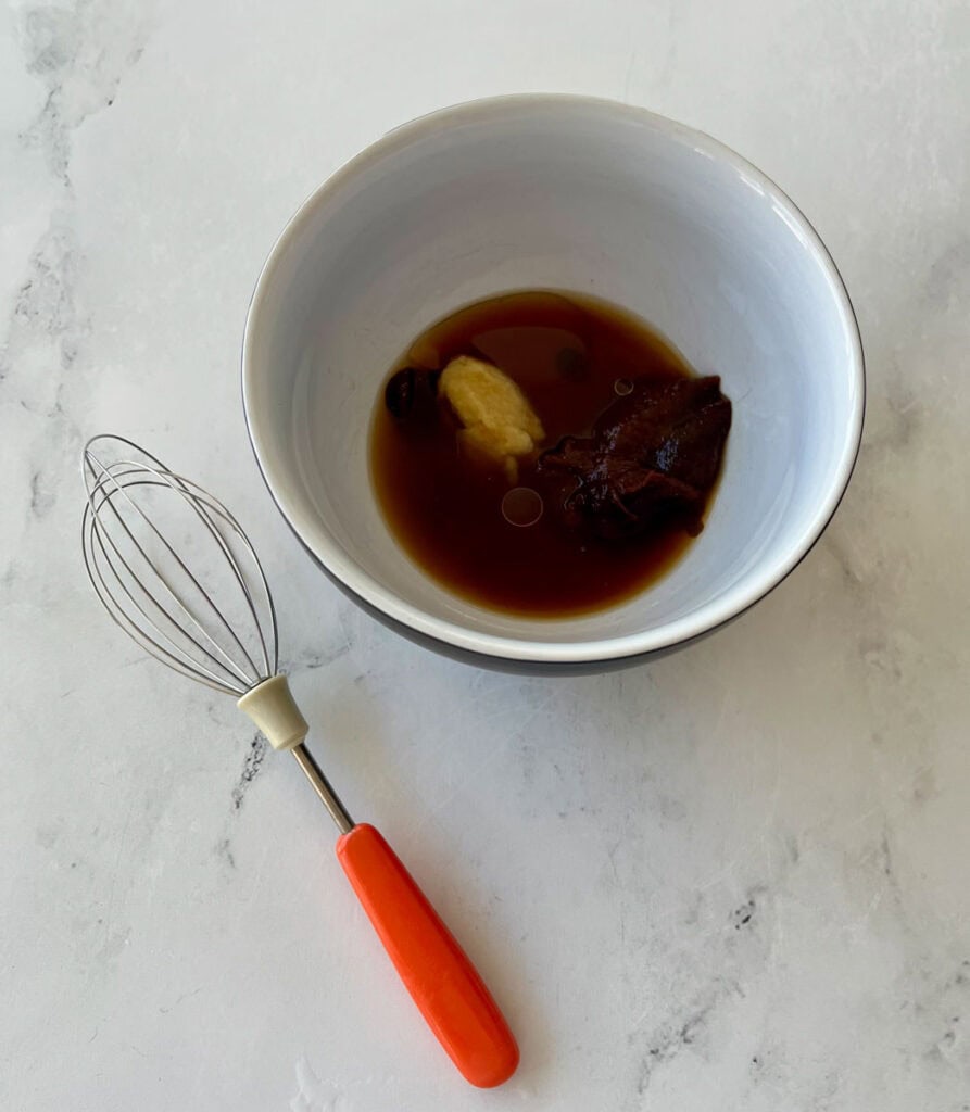 There is a white bowl with a dark sauce in it. There is a whisk with a red handle laying next to the bowl. Items are on a white surface.