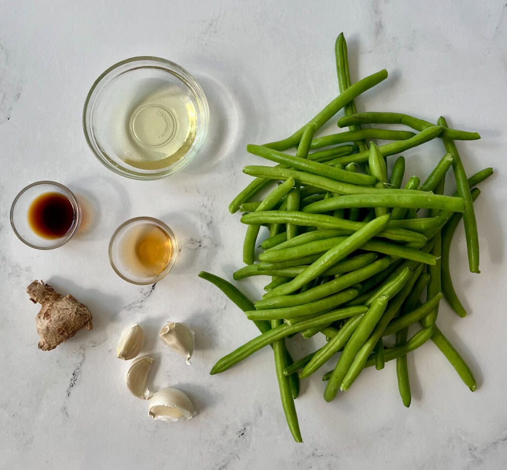 There is a mound of fresh green beans, cloves of garlic, a piece of ginger root, three small clear dishes, one with sesame oil, one with soy sauce, and one with rice wine in it. Items are on a white surface.