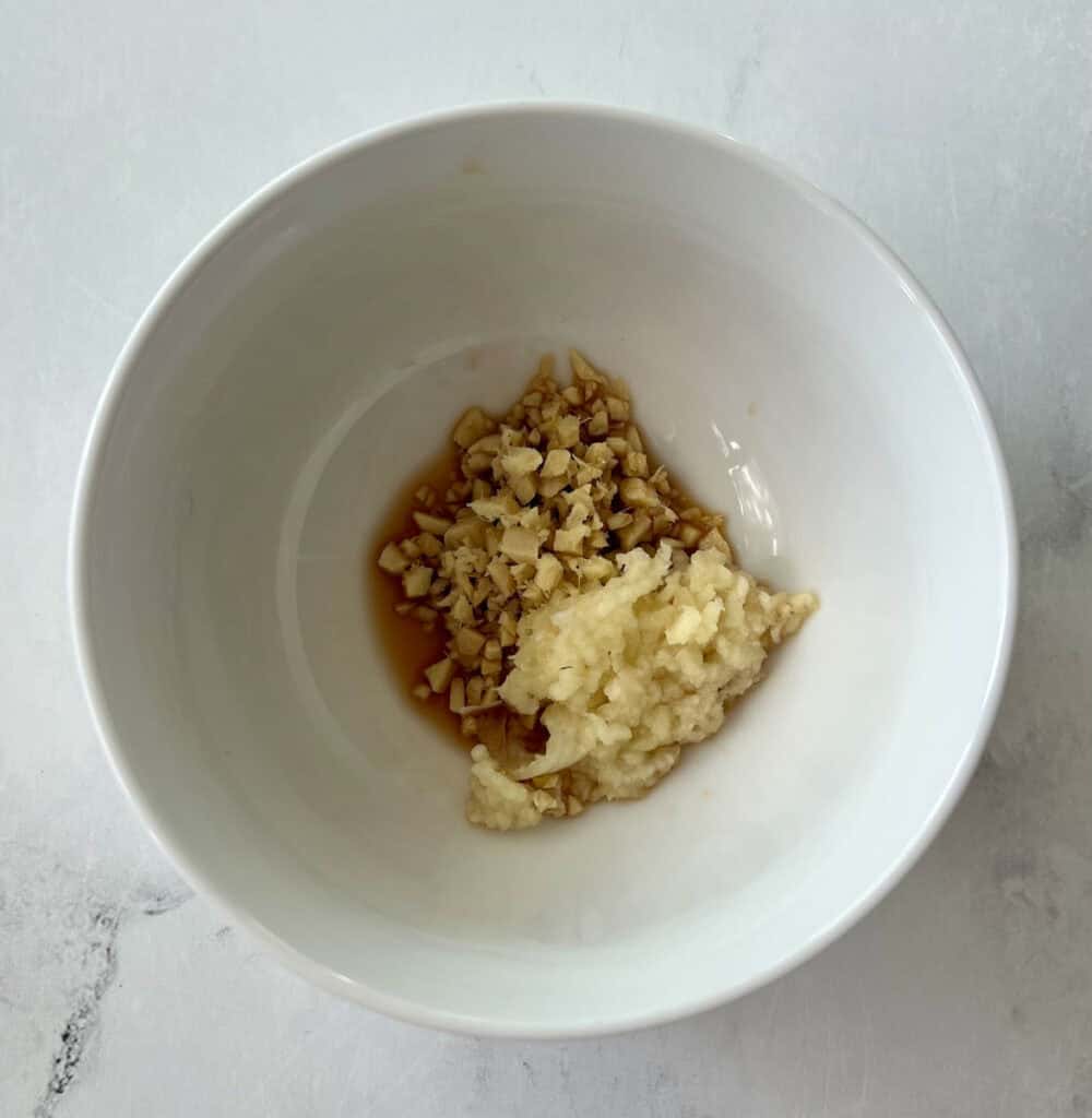 Small white mixing bowl with chopped ginger, minced garlic, rice wine, soy sauce, and sesame oil in it. Bowl is on a white surface.