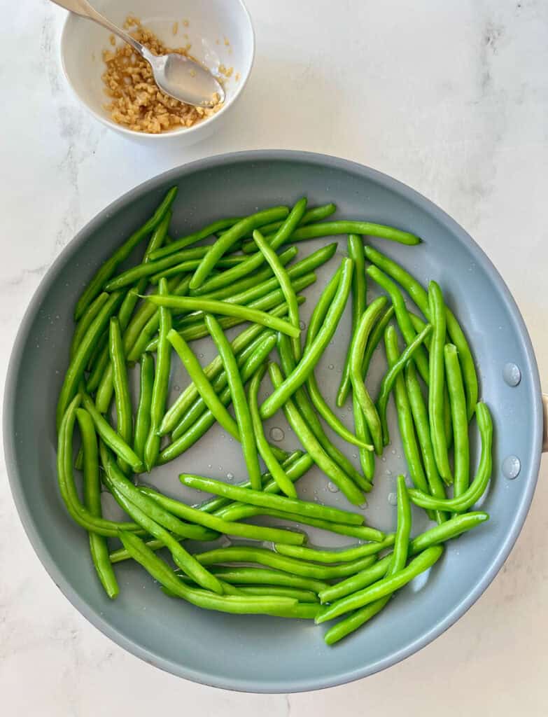 Large gray fry pan with green beans in it. There is a small white bowl with a ginger and garlic mixture in it. There is a silver spoon resting in the bowl. Items are on a white surface.