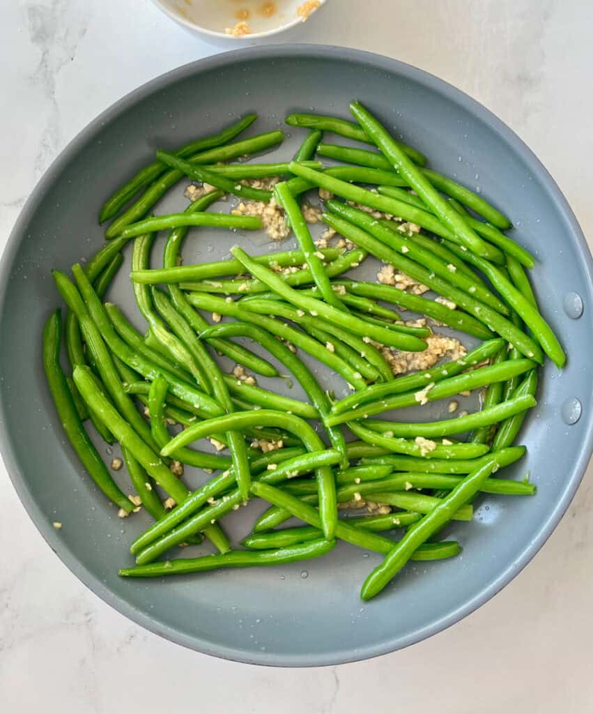 Large gray fry pan with green beans tossed in a ginger and garlic mixture. Pan is on a white surface.
