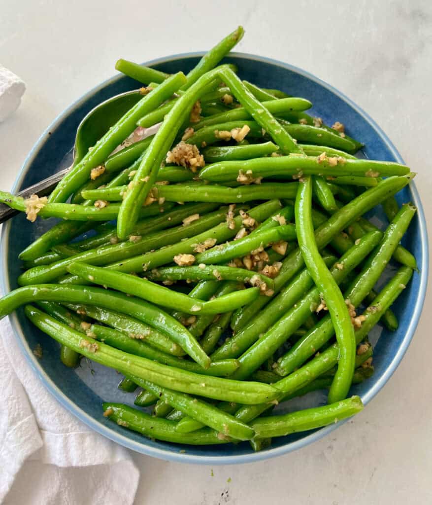 There is a large shallow light blue serving dish with cooked green beans tossed in a ginger and garlic mixture. There is a large spoon resting in the dish. There is a white cloth napkin. Items are on a white surface.