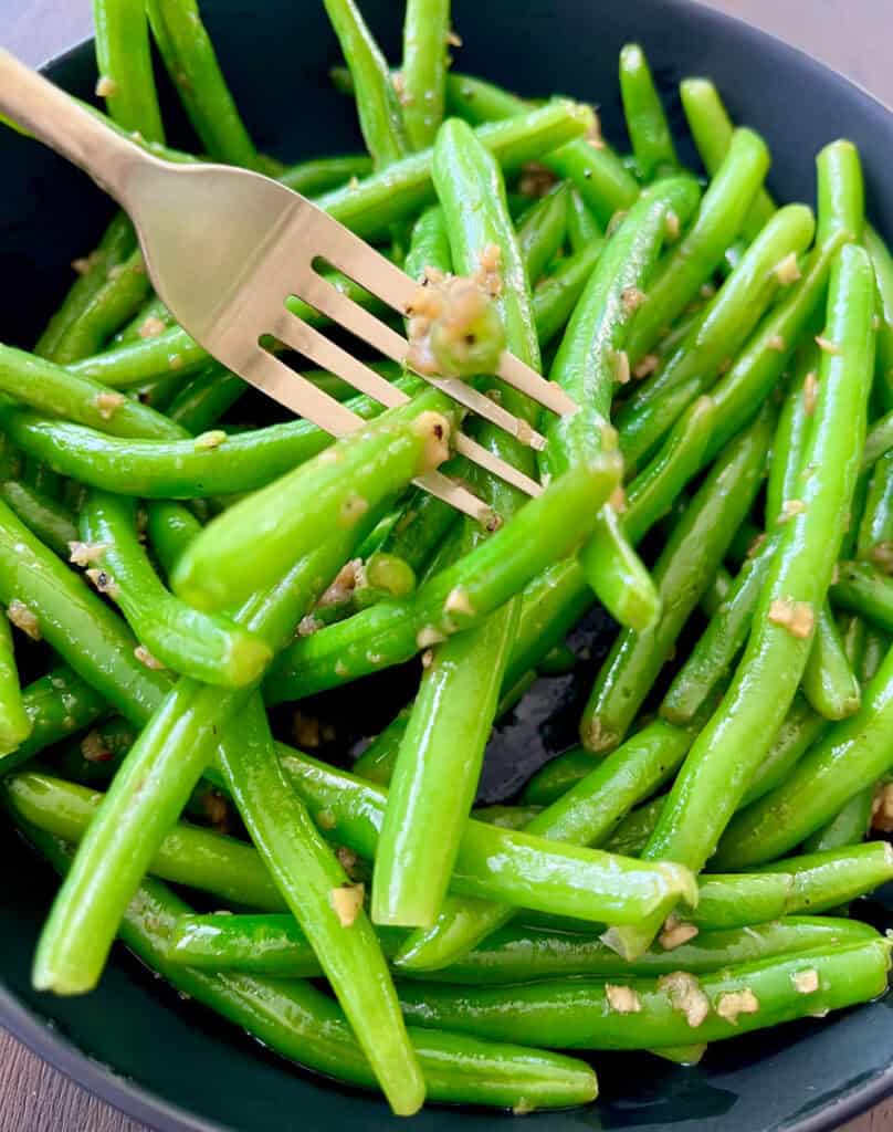 Large black serving bowl with green beans tossed in a ginger and garlic mixture. There is a fork with beans on it in the bowl.