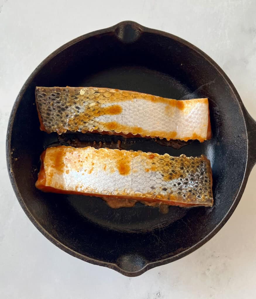 Black cast iron skillet with two salmon filets cooking, skin side up. Skillet is on a white surface.