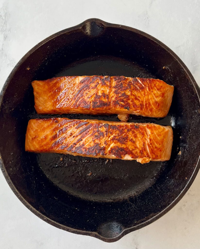 Black cast iron skillet with two salmon filets cooking. Skillet is on a white surface.
