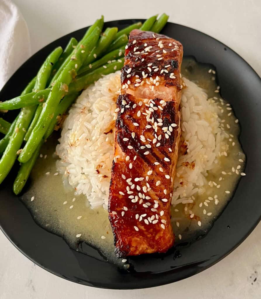 There is a black plate with a mound of white rice in a pool of sauce, with a piece of salmon on top of the rice. There are sesame seeds sprinkled over the top. There are green beans on the plate. Plate is on a white surface.