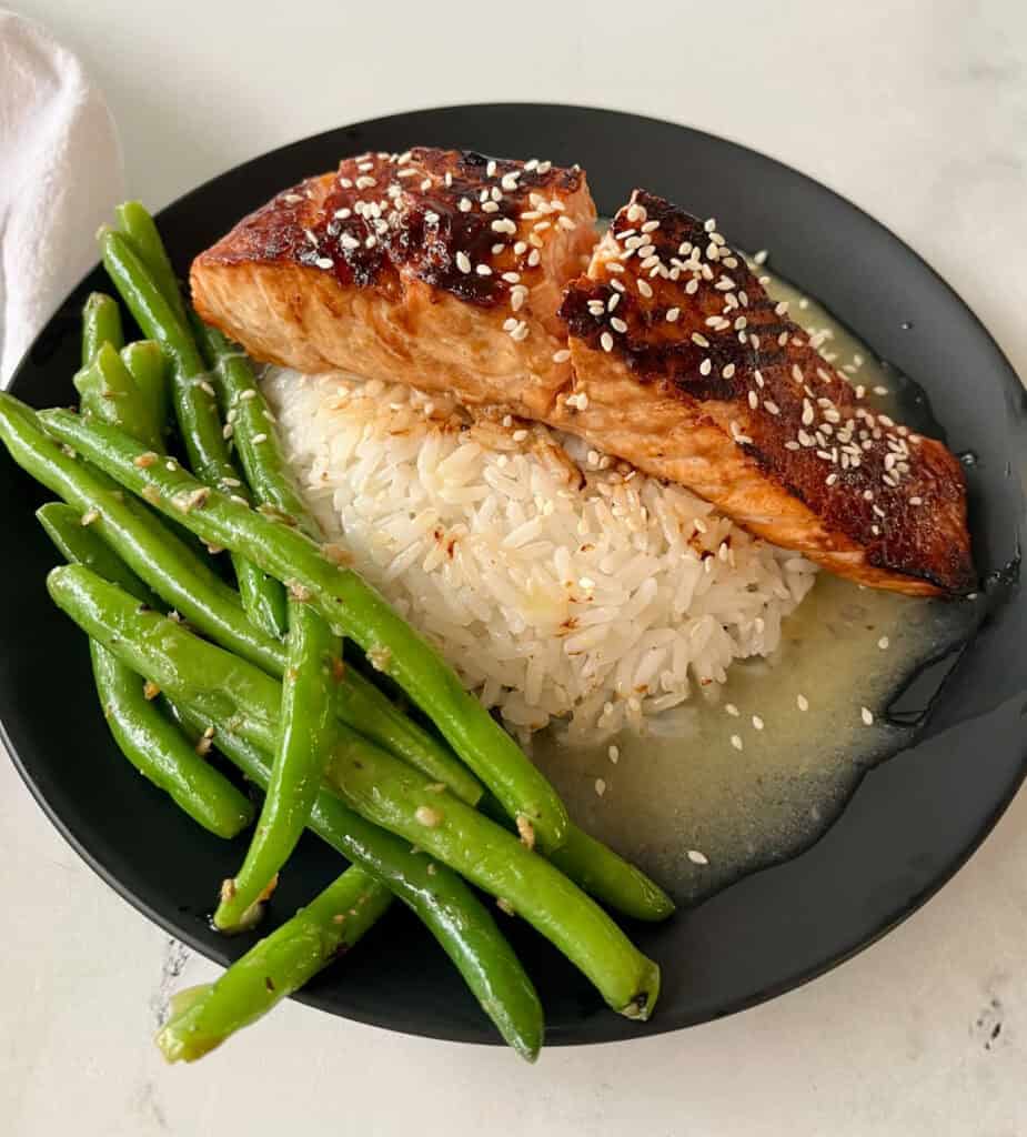 There is a black plate with a mound of white rice in a pool of sauce, with a piece of salmon on top of the rice. There are sesame seeds sprinkled over the top. There are green beans on the plate. Plate is on a white surface.