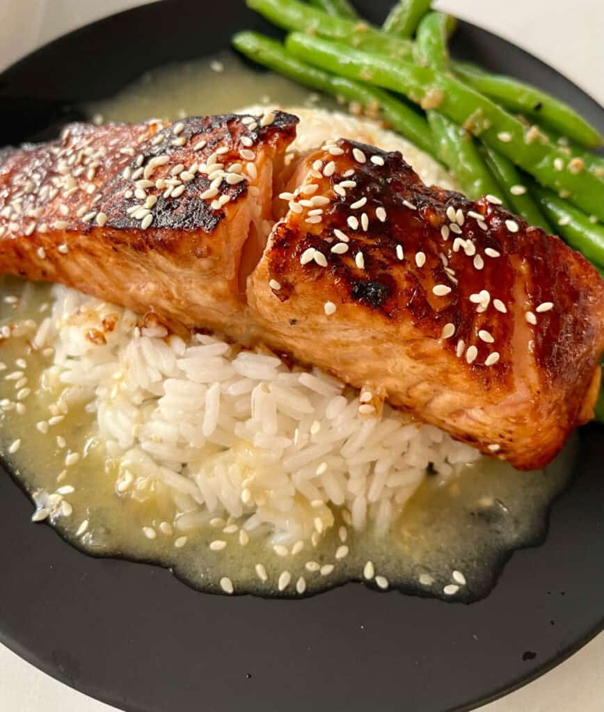There is a black plate with a mound of white rice in a pool of sauce, with a piece of salmon on top of the rice. There are sesame seeds sprinkled over the top. There are green beans on the plate.