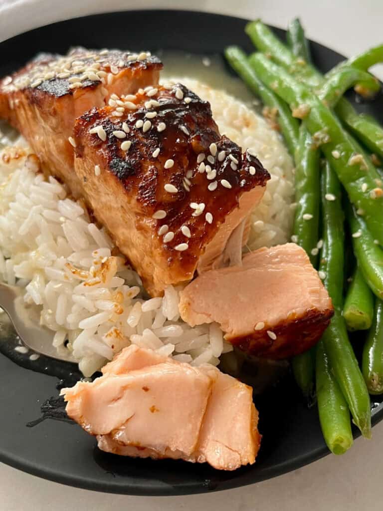 There is a black plate with a mound of white rice in a pool of sauce, with a piece of salmon on top of the rice. There are sesame seeds sprinkled over the top. There are green beans on the plate. Plate is on a white surface.