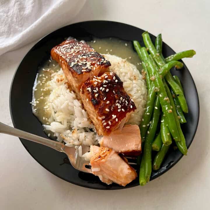 There is a black plate with a mound of white rice in a pool of sauce, with a piece of salmon on top of the rice. There are sesame seeds sprinkled over the top. There is a fork with a piece of salmon on it. There are green beans on the plate. There is a white cloth napkin. Items are on a white surface.
