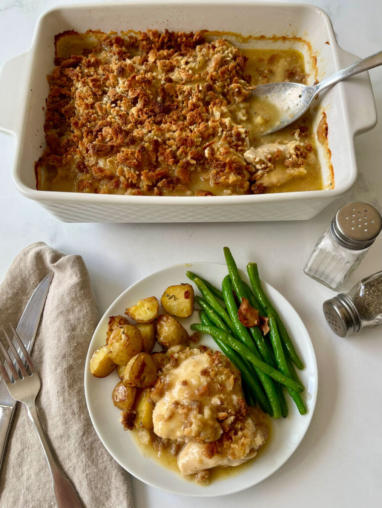 There is a white plate with small roasted potatoes, green beans and a serving of baked chicken in a sauce and baked bread crumbs. There is a large white rectangular baking dish with pieces of baked chicken in a sauce with a crispy bread crumb topping. There is a knife and fork laying on a light brown cloth napkin and a salt and pepper shaker. Items are on a white surface.