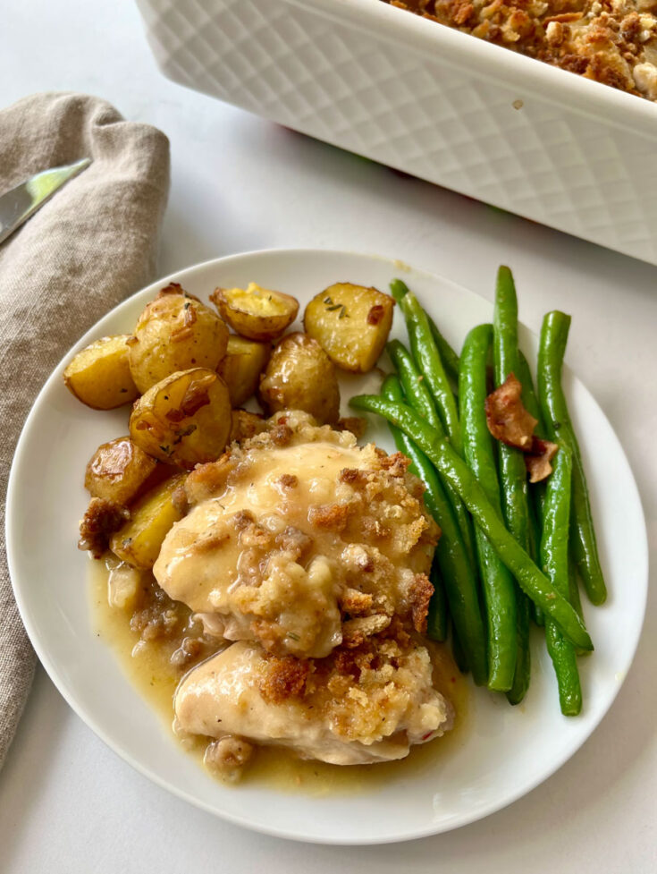 There is a white plate with small roasted potatoes, green beans and a serving of baked chicken in a sauce and baked bread crumbs. There is a white casserole dish and a light brown cloth napkin. Items are on a white surface.