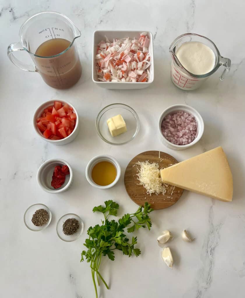 Various size bowls with ingredients in them: chopped crabmeat, chopped shallots, chopped tomatoes, butter, ground pepper, dry thyme, tomato paste, and sherry. There are two glass measuring cups. One with chicken broth and one with heavy cream. There are sprigs of parsley, cloves of garlic and a wedge of parmesan cheese on a small wooden board. Items are on a white surface.