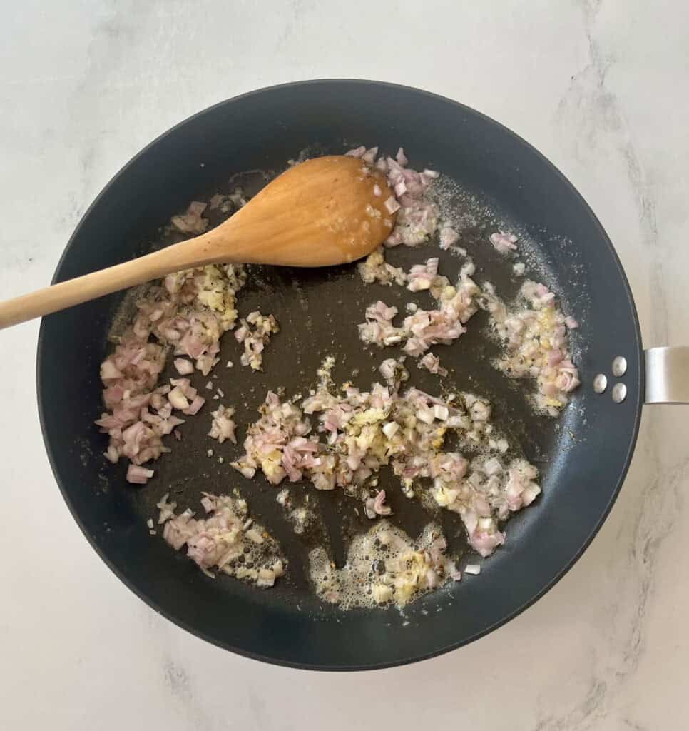 There is a large gray skillet with chopped shallots and minced garlic cooking in butter. There is a wooden spoon resting in the skillet. Skillet is on a white surface.