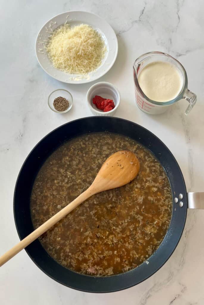 There is a large gray skillet with chopped shallots, minced garlic and chicken broth cooking. There is a wooden spoon resting in the skillet. There is a clear glass measuring cup with heavy cream in it, a plate with grated parmesan cheese on it, two small bowls, one with dry herbs in it and the other with tomato paste in it in the background. Items are on a white surface.