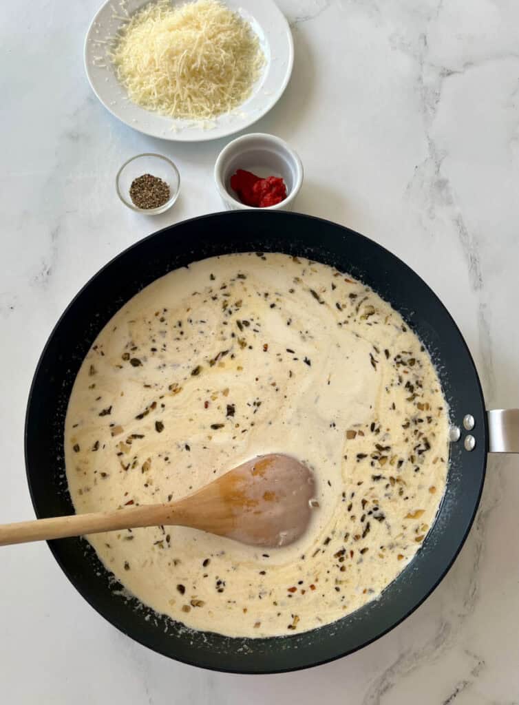 There is a large gray skillet with chopped shallots, minced garlic, chicken broth, and heavy cream cooking. There is a wooden spoon resting in the skillet. There is a plate with grated parmesan cheese on it, two small bowls, one with dry herbs in it and the other with tomato paste in it in the background. Items are on a white surface.