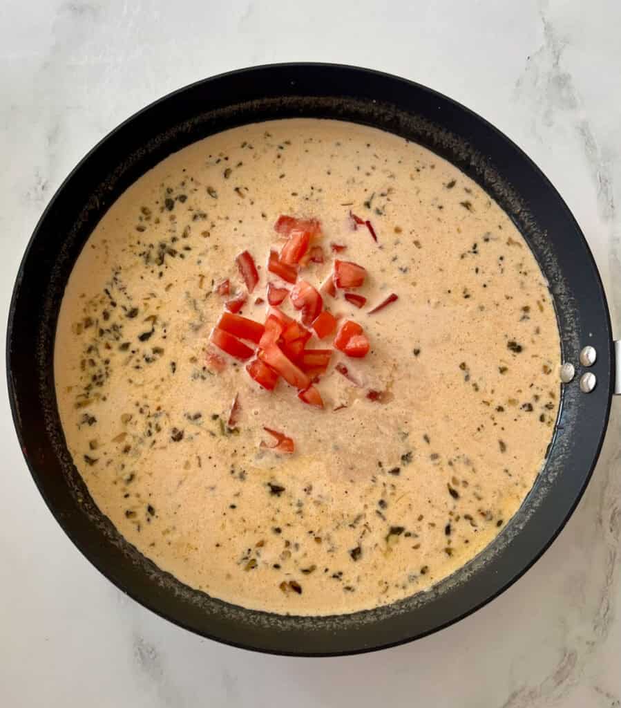 There is a large gray skillet with a cream sauce with tomatoes, cheese and herbs cooking in it. Skillet is on a white surface.