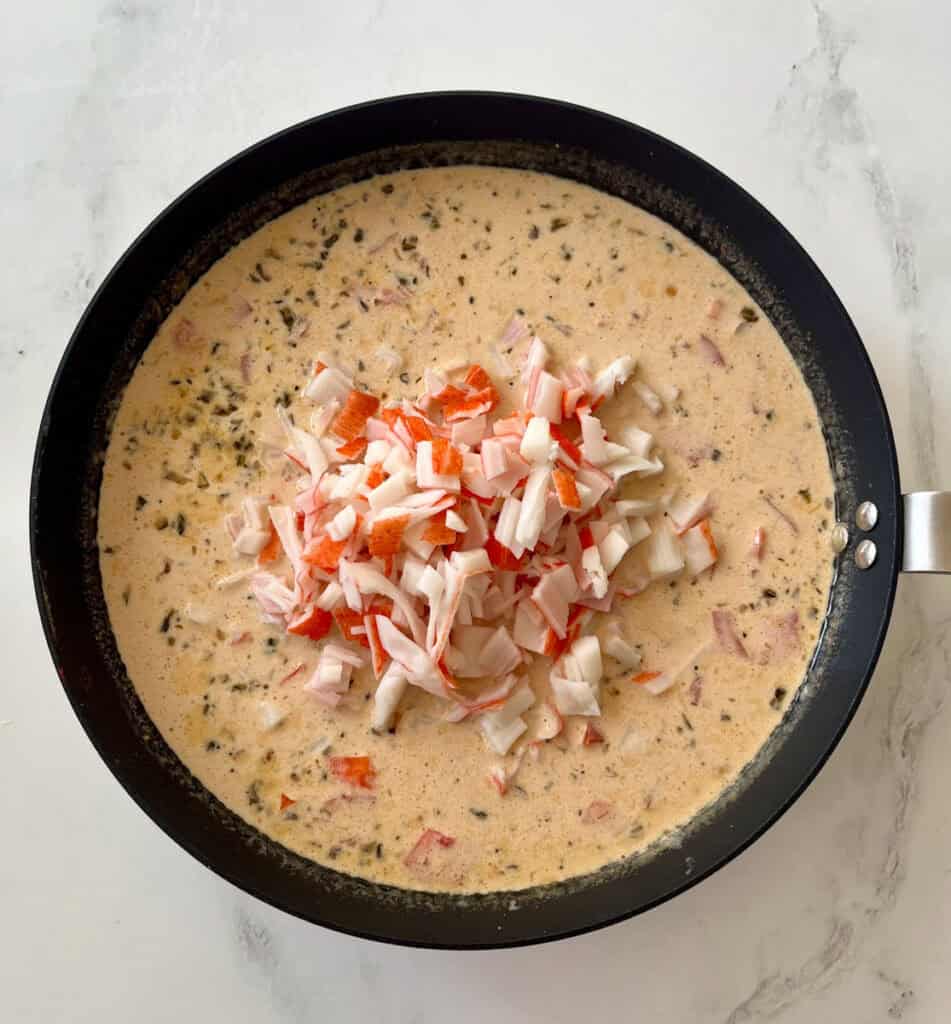 Large gray skillet with a cream sauce with tomatoes, cheese and herbs cooking in it. There is a mound of chopped crabmeat in the center of the skillet. Skillet is on a white surface.
