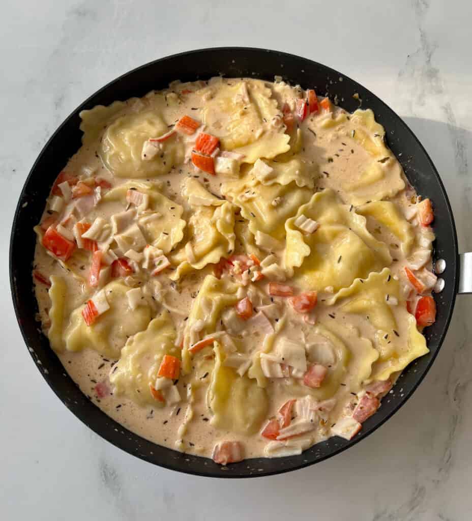 Large dark skillet with ravioli/pasta and a cream sauce that has chopped crabmeat and chopped tomatoes. Skillet is on a white surface.