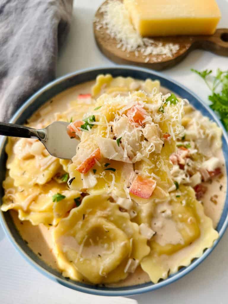 There is a blue shallow bowl with ravioli/pasta and a cream sauce that has chopped crabmeat and chopped tomatoes. There is chopped fresh parsley sprinkled over the top. There is a fork resting in the bowl. In the background there is a gray cloth napkin, sprigs of fresh parsley, and a block of parmesan cheese. Items are on a white surface.