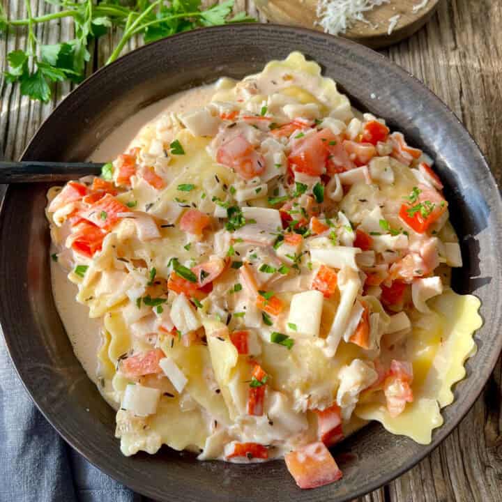 There is a dark bowl with ravioli/pasta and a cream sauce that has chopped crabmeat and chopped tomatoes. There is chopped fresh parsley sprinkled over the top. There is a spoon resting in the bowl. In the background there is a gray cloth napkin, sprigs of fresh parsley, and a block of parmesan cheese. Items are on a wood surface.