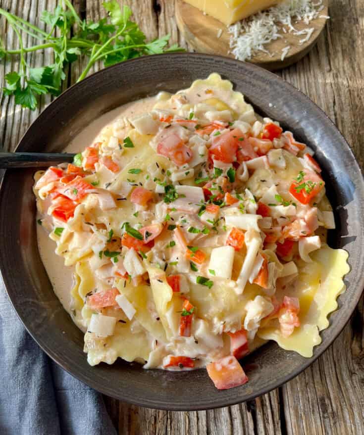 There is a dark bowl with ravioli/pasta and a cream sauce that has chopped crabmeat and chopped tomatoes. There is chopped fresh parsley sprinkled over the top. There is a spoon resting in the bowl. In the background there is a gray cloth napkin, sprigs of fresh parsley, and a block of parmesan cheese. Items are on a wood surface.