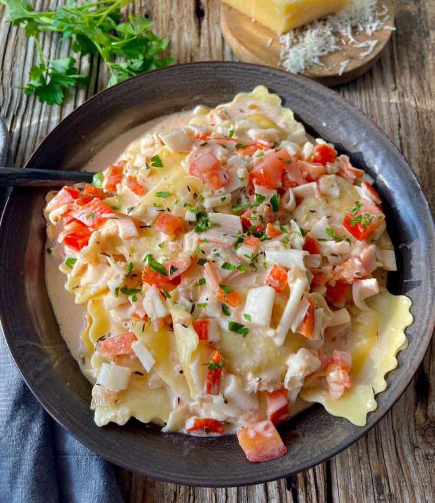 There is a dark bowl with ravioli/pasta and a cream sauce that has chopped crabmeat and chopped tomatoes. There is chopped fresh parsley sprinkled over the top. There is a spoon resting in the bowl. In the background there is a gray cloth napkin, sprigs of fresh parsley, and a block of parmesan cheese. Items are on a wood surface.