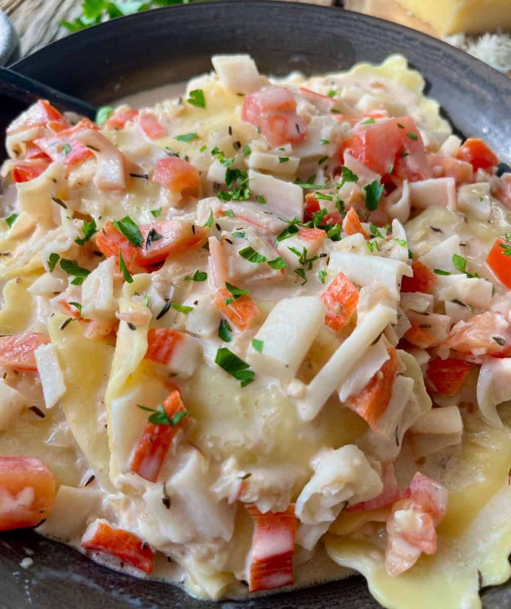 There is a dark bowl with ravioli/pasta and a cream sauce that has chopped crabmeat and chopped tomatoes. There is chopped fresh parsley sprinkled over the top.